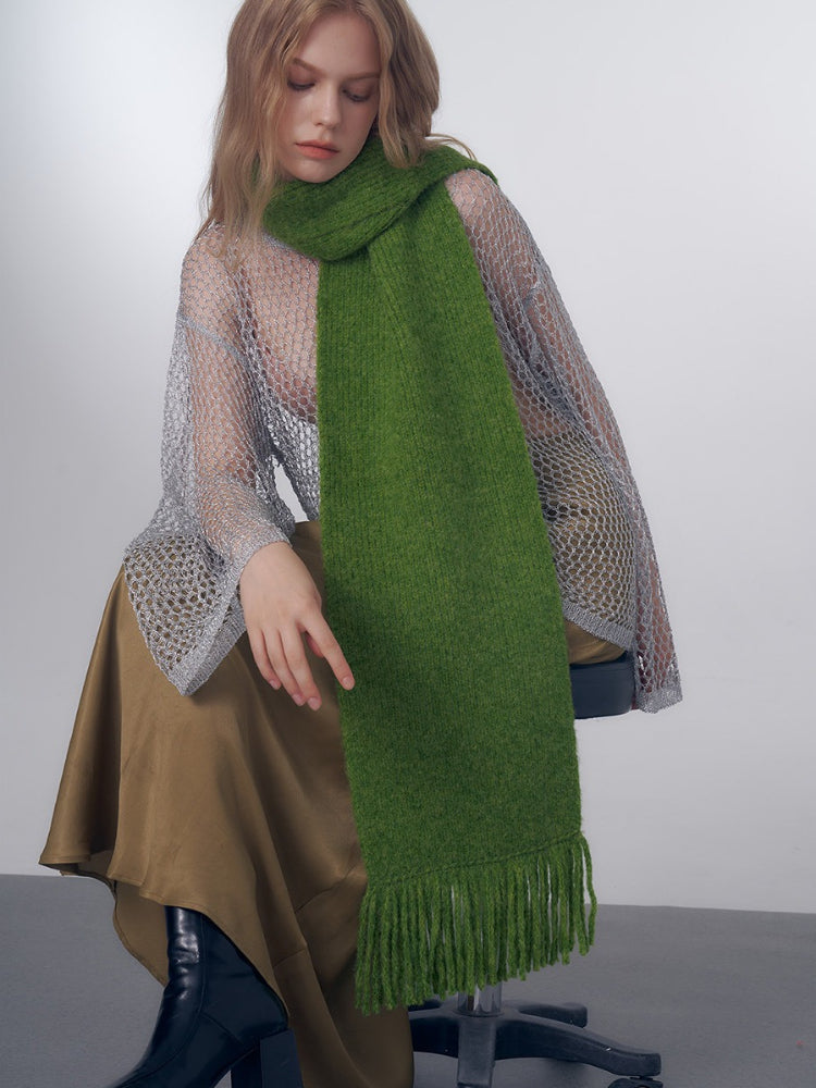 Model wearing a green knitted scarf with fringe over a sheer silver top and a brown skirt.