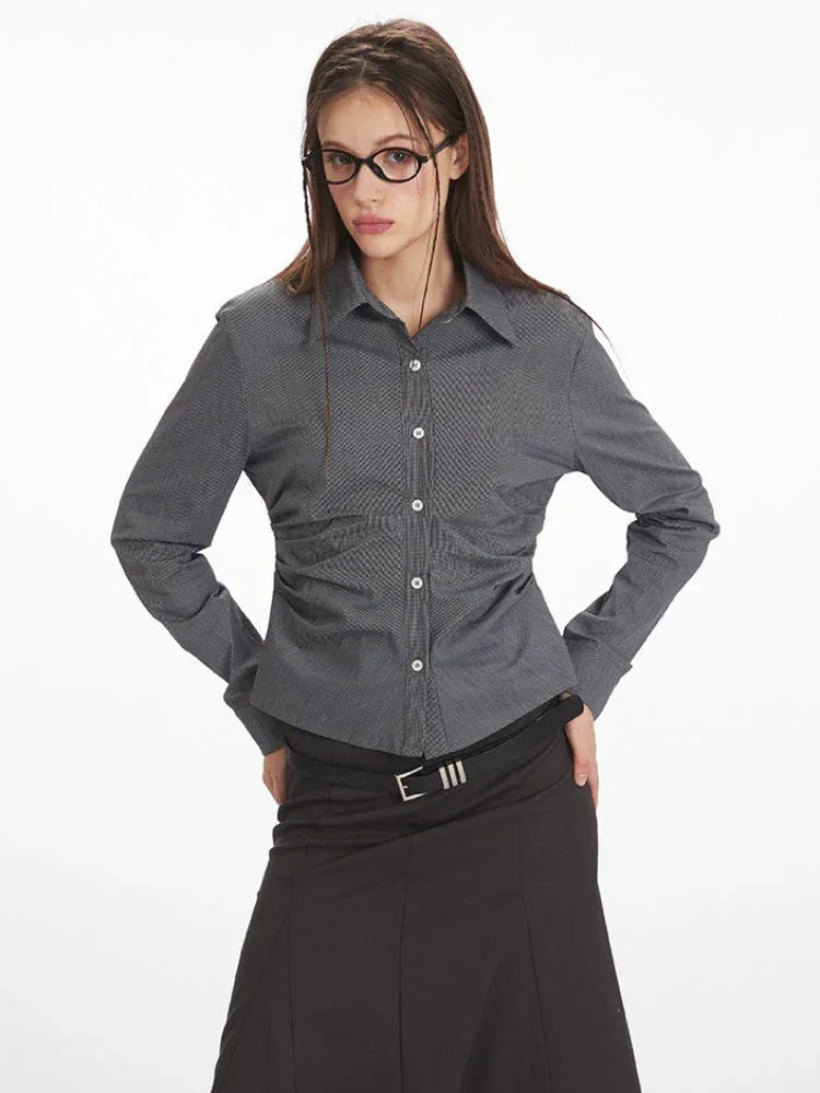 Close-up of a woman wearing a grey long-sleeved button-down shirt with ruching detail and a black belt over a black skirt.