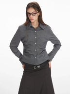 Close-up of a woman wearing a grey long-sleeved button-down shirt with ruching detail and a black belt over a black skirt.