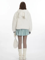 Back view of a model wearing a white fuzzy hooded jacket with bunny ears, a light blue pleated skirt, grey tights, and white leg warmers. She is holding a white fuzzy purse.