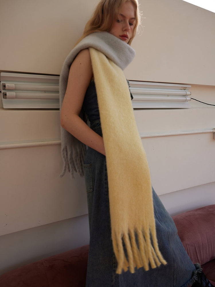 Woman wearing a two-tone yellow and gray fringed scarf draped over a denim dress, leaning against a wall.