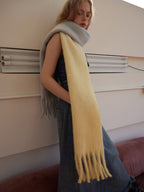Woman wearing a two-tone yellow and gray fringed scarf draped over a denim dress, leaning against a wall.