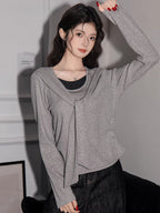 Woman wearing a grey long-sleeved top with a draped overlay and black pants, with her arm raised.
