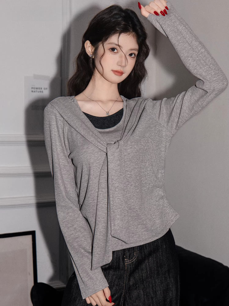 Woman wearing a grey long-sleeved top with a draped overlay and black pants, with her arm raised.