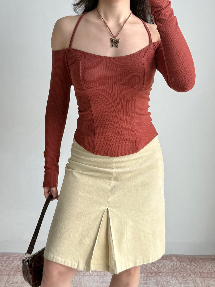 Full body shot of a woman wearing a rust-colored ribbed knit top with off-the-shoulder long sleeves and a halter neckline, paired with a cream-colored skirt and holding a dark brown handbag.