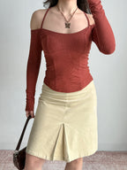 Full body shot of a woman wearing a rust-colored ribbed knit top with off-the-shoulder long sleeves and a halter neckline, paired with a cream-colored skirt and holding a dark brown handbag.