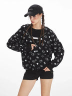 Front view of a model wearing a black zip-up hoodie with an all-over white Hello Kitty and heart graphic print, black t-shirt, black shorts, black knee-high socks, and red sneakers. The model is wearing a black baseball cap and has her hair in braids, with one hand in the hoodie pocket.