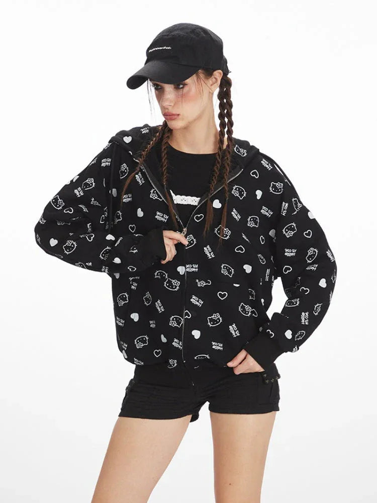 Front view of a model wearing a black zip-up hoodie with an all-over white Hello Kitty and heart graphic print, black t-shirt, black shorts, black knee-high socks, and red sneakers. The model is wearing a black baseball cap and has her hair in braids, with one hand in the hoodie pocket.