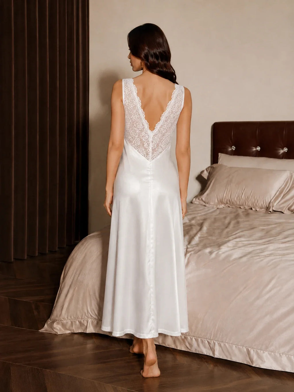 Back view of a woman wearing a white satin and lace maxi slip dress, showing the V-back design, in a bedroom setting.