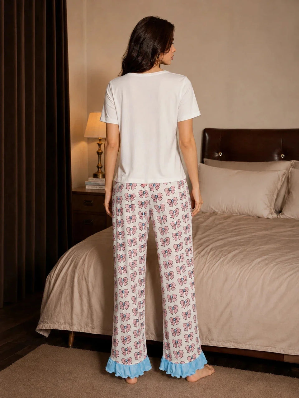 Back view of a woman wearing a white short-sleeve pajama top and white pajama pants with a pink bow print and blue ruffle hem, standing in a bedroom. Secondary image