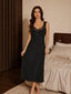Front view of a woman wearing a black rib-knit midi nightdress with lace trim and a bow detail, standing in a bedroom.