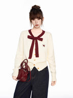 Woman holding a burgundy handbag while wearing a cream-colored cardigan with a burgundy bow and trim and dark wash wide-leg jeans.
