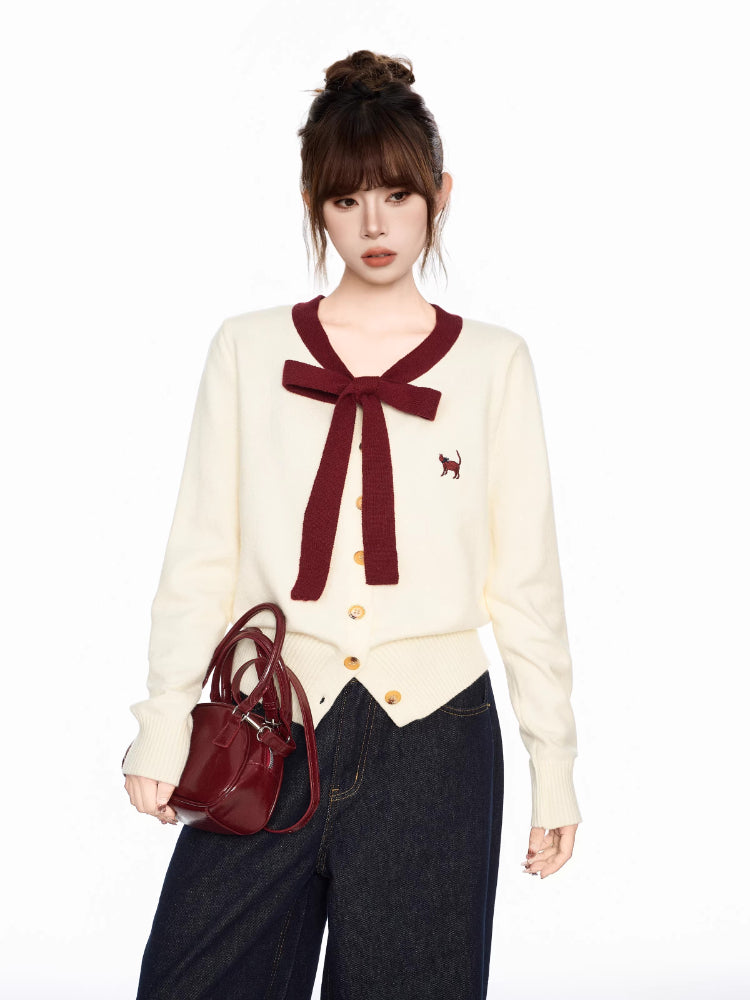 Woman holding a burgundy handbag while wearing a cream-colored cardigan with a burgundy bow and trim and dark wash wide-leg jeans.