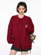 Woman wearing a red knit cardigan with tie neck and buttons, grey pleated skirt, brown crossbody bag, and white leg warmers.