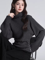 Woman sitting and wearing a dark grey turtleneck sweater with batwing sleeves and a cinched button detail at the waist.