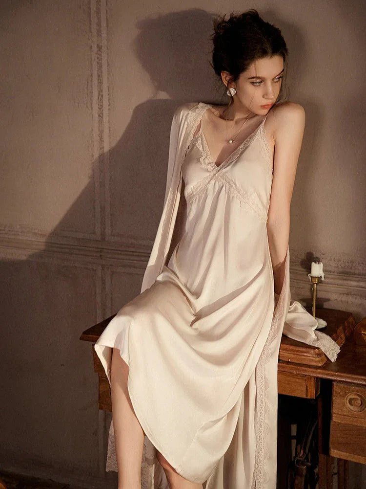 Woman wearing a cream satin slip dress with lace trim and a matching robe, leaning on a wooden table.