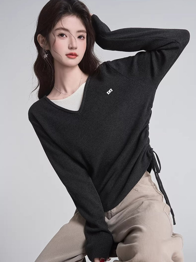 Woman wearing a dark grey v-neck sweater with a white layer underneath and tan pants, posing with her arm raised.