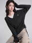 Woman wearing a dark grey v-neck sweater with a white layer underneath and tan pants, posing with her arm raised.