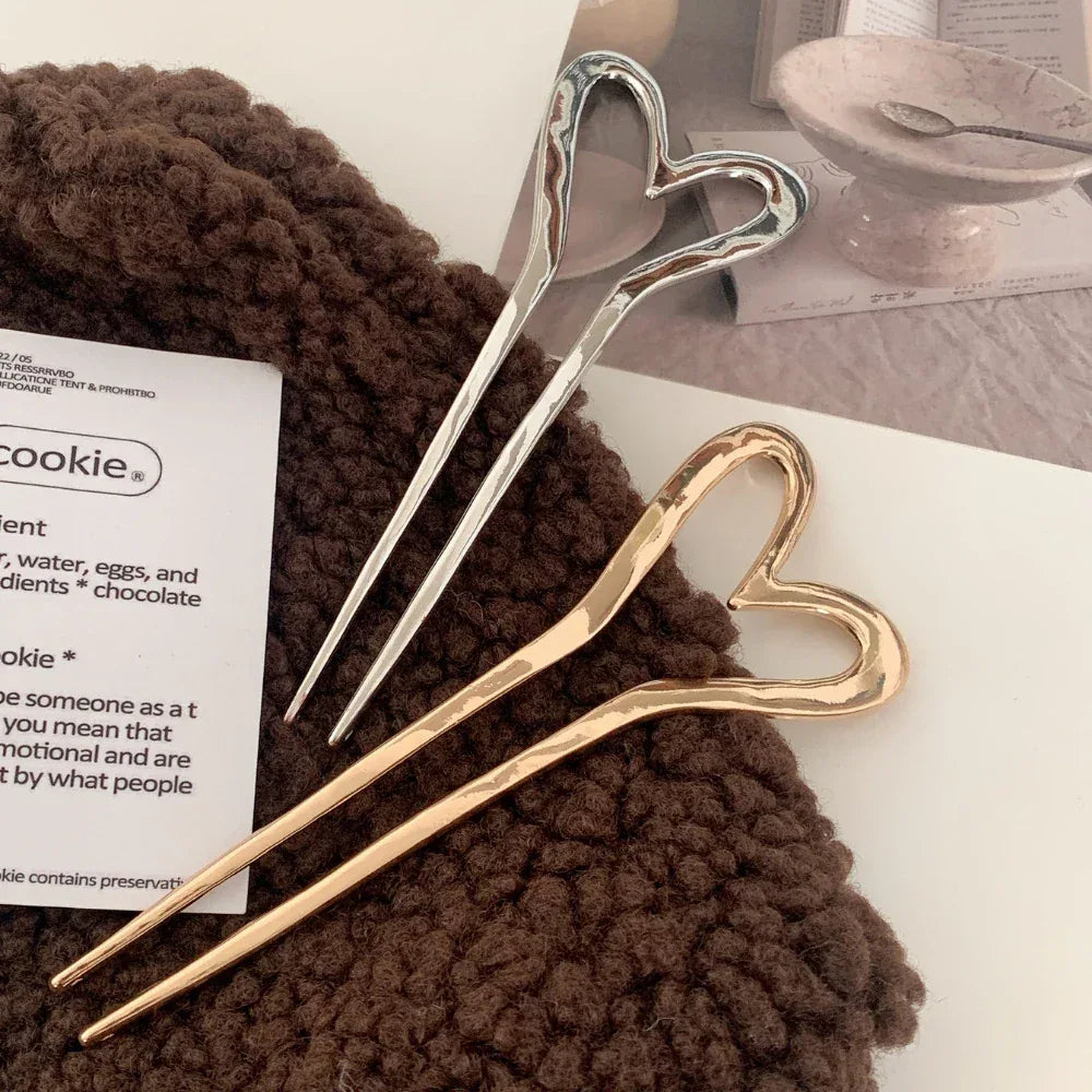 Two heart-shaped hairpins, one gold-tone and one silver-tone, resting on a textured brown fabric.