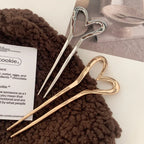 Two heart-shaped hairpins, one gold-tone and one silver-tone, resting on a textured brown fabric.