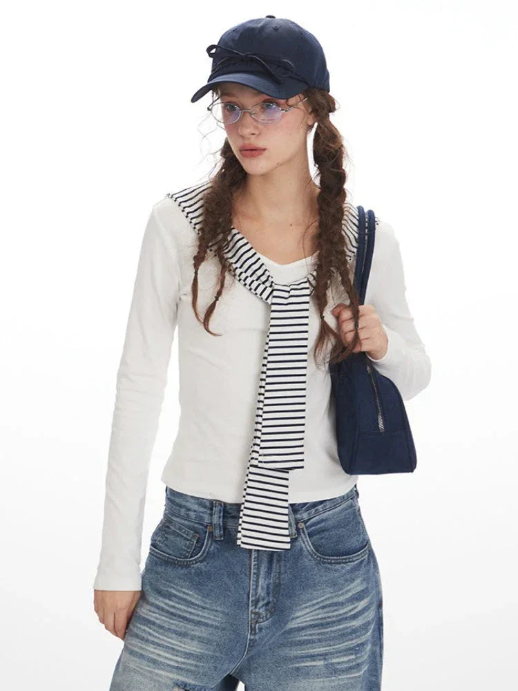 Close-up of a model wearing a white long-sleeved top with striped trim and wide-leg blue jeans, holding a navy handbag and wearing a navy baseball cap.