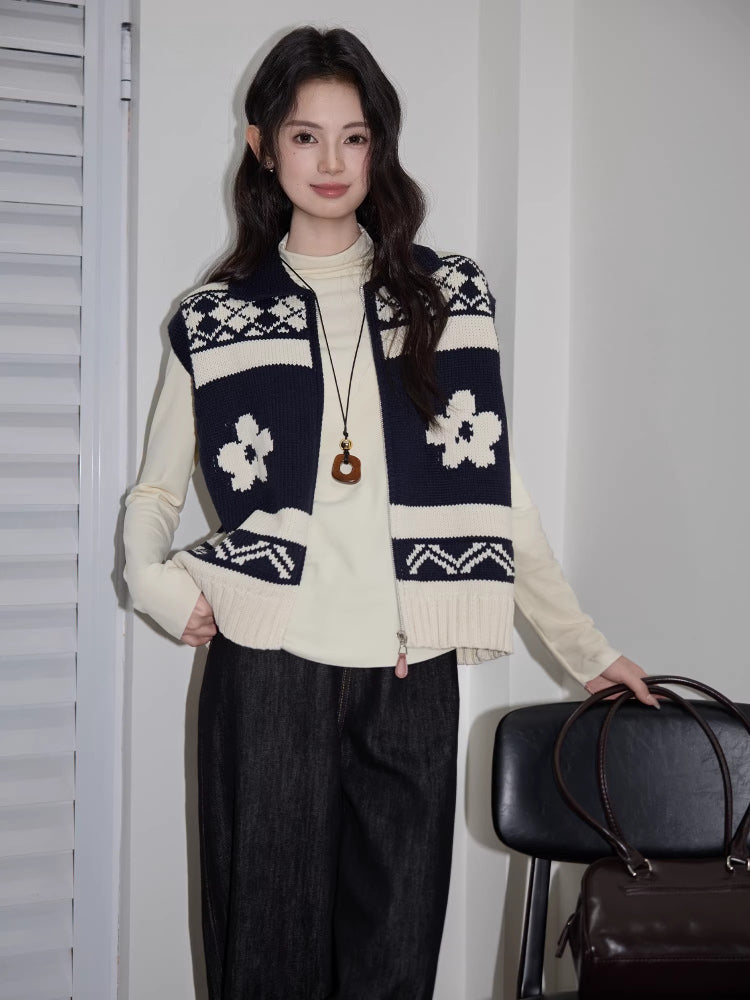 Woman wearing a navy and cream floral and geometric knit vest over a cream long-sleeve top and dark wash wide-leg jeans, standing and holding a handbag.