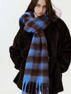A woman stands with her hands in the pockets of a dark, textured coat, wearing a large, blue and brown plaid scarf with fringe.