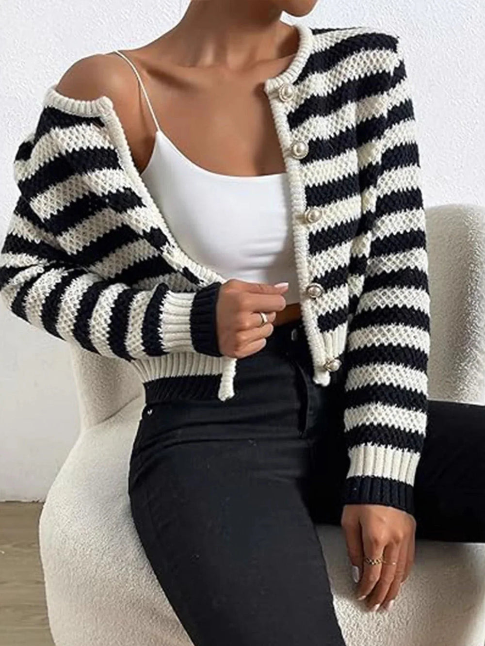Woman wearing a black and white striped cropped knit cardigan over a white camisole and black jeans, showing the front of the cardigan with one side open.