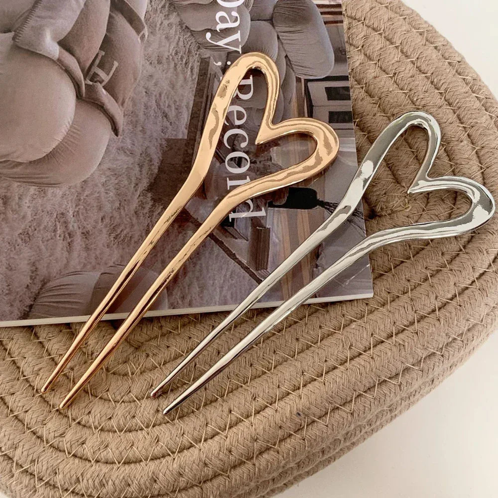 Three heart-shaped hairpins, two gold-tone and one silver-tone, arranged on a woven mat over a magazine.