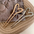 Three heart-shaped hairpins, two gold-tone and one silver-tone, arranged on a woven mat over a magazine.