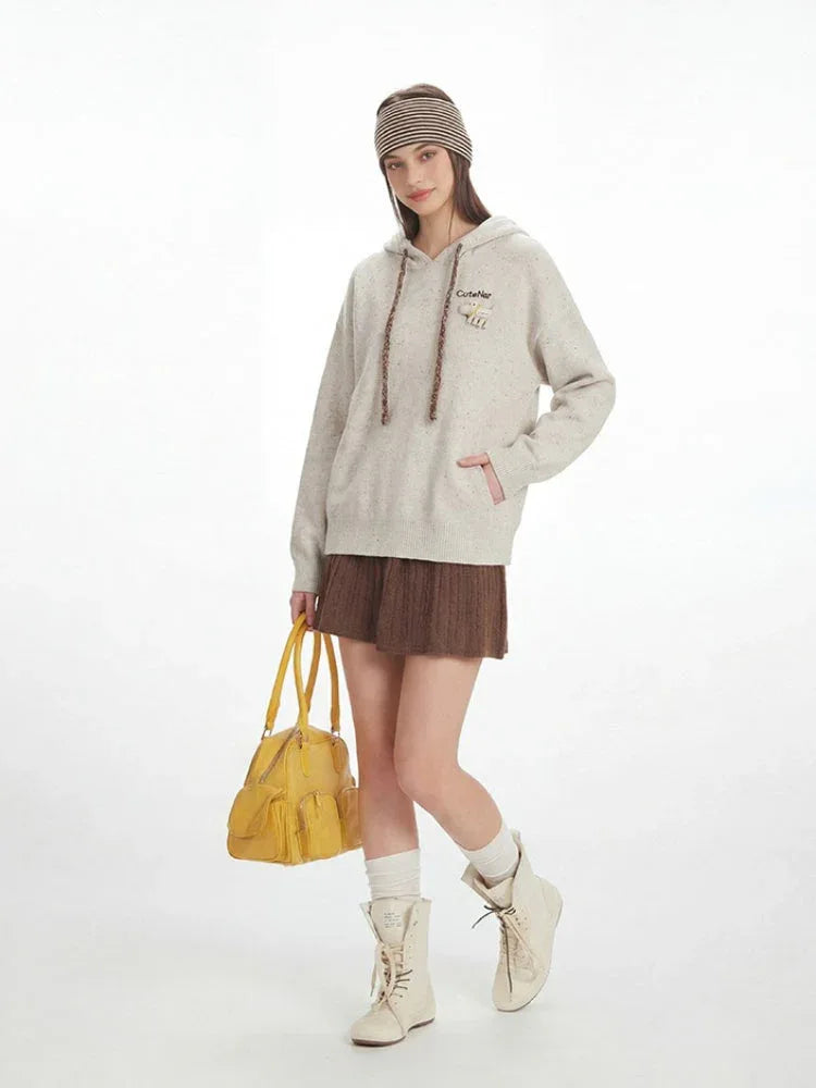 Full-body shot of a model in a casual outfit featuring a speckled knit hoodie, brown pleated skirt, white socks, and cream boots, with a yellow handbag.