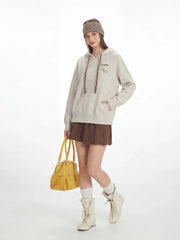 Full-body shot of a model in a casual outfit featuring a speckled knit hoodie, brown pleated skirt, white socks, and cream boots, with a yellow handbag.