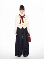 Full-length shot of a woman in a cream-colored cardigan with a burgundy bow and trim, dark wash wide-leg jeans, and a burgundy handbag.