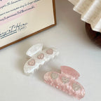 Two marble claw clips, one pink and one white, each with three heart-shaped embellishments with rhinestones, displayed on a white surface next to lace and a wooden bowl