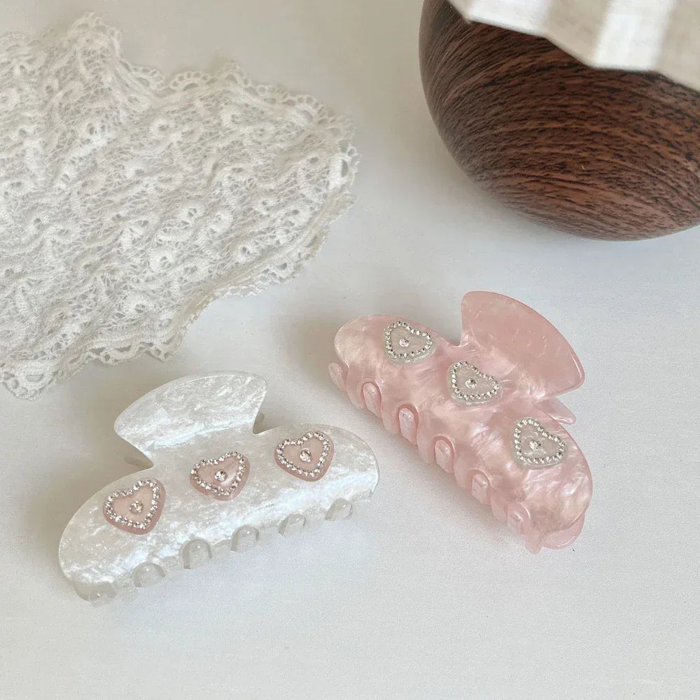 Two marble claw clips, one pink and one white, each with three heart-shaped embellishments with rhinestones, displayed on a white surface next to lace and a wooden bowl