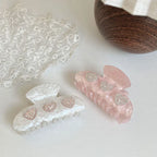 Two marble claw clips, one pink and one white, each with three heart-shaped embellishments with rhinestones, displayed on a white surface next to lace and a wooden bowl