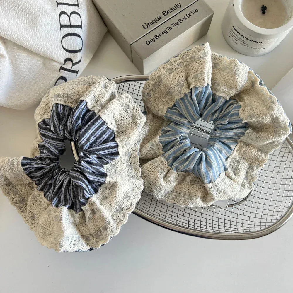 Two scrunchies are placed on a wire basket, one with dark blue stripes and the other with light blue stripes, both adorned with a ruffled lace overlay.