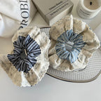 Two scrunchies are placed on a wire basket, one with dark blue stripes and the other with light blue stripes, both adorned with a ruffled lace overlay.