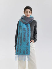 A woman models a blue and gray knit scarf with text print and fringe detail, worn over a gray sweater and cream pants.