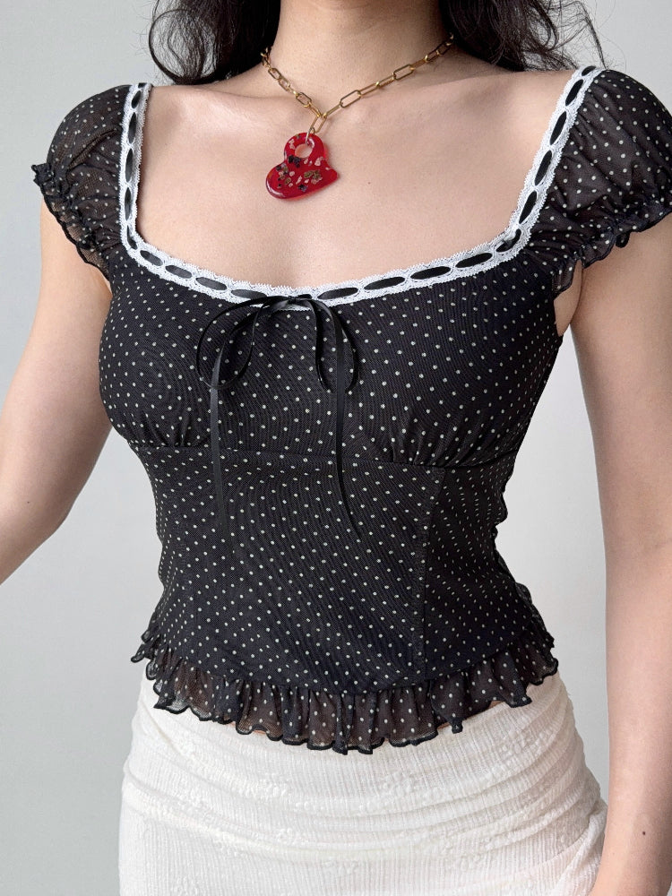 Side view of a woman wearing a black and white polka dot mesh top with a square neckline and ruffled sleeves.