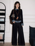 Model standing, showcasing a layered black top with checkered collar and cuffs, dark wash wide-leg jeans, and a brown shoulder bag, with a bookshelf in the background.