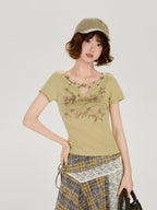 Model with arms crossed, wearing a light green short-sleeve t-shirt with a cutout neckline and star details, paired with an asymmetrical plaid skirt and a khaki baseball cap, holding a brown handbag.