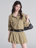 Woman wearing a tan button-down shirt dress with a black studded belt and a black shoulder bag.