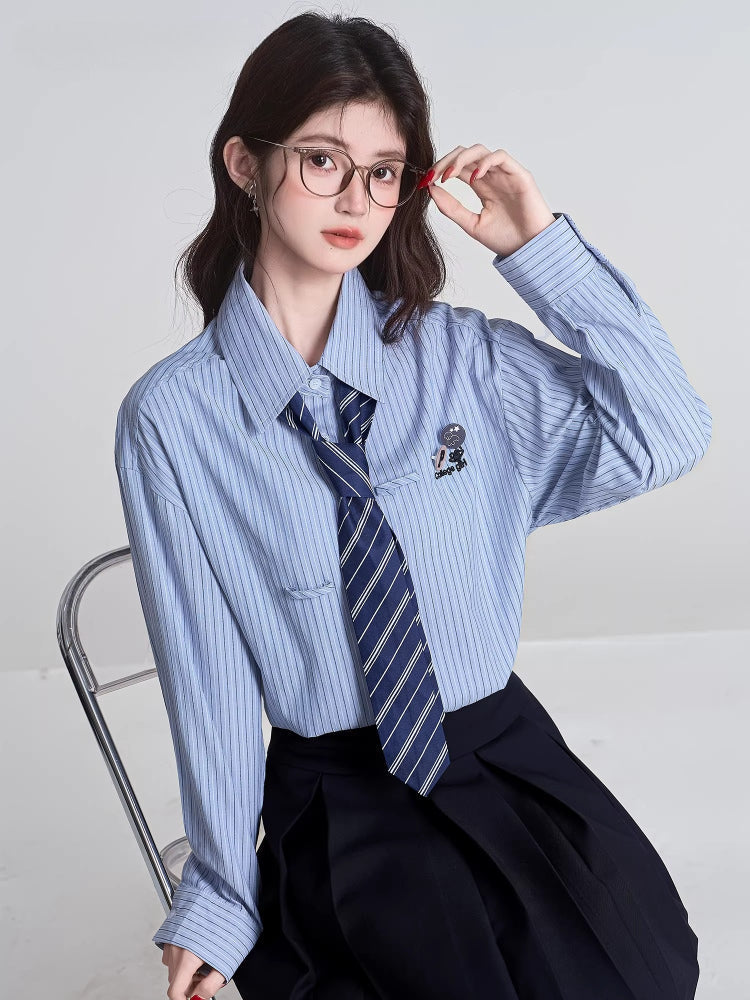 Model wearing a light blue striped button-down shirt with a navy striped tie and a black pleated skirt, adjusting glasses.