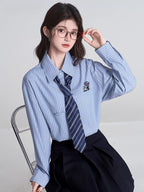 Model wearing a light blue striped button-down shirt with a navy striped tie and a black pleated skirt, adjusting glasses.
