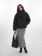 Full-length view of a woman in a black puffer jacket, grey skirt, and black boots, carrying a tote bag.