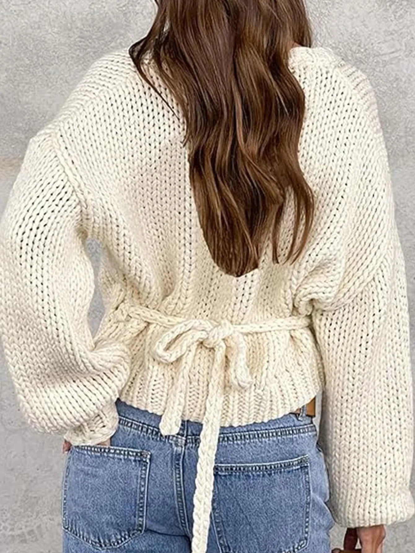 Back view of a woman wearing a cream chunky knit wrap sweater tied at the waist with light wash jeans.
