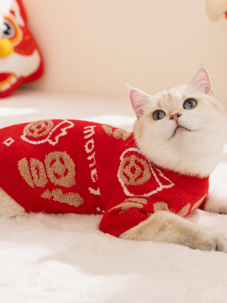 A white cat lying down, wearing a red knit sweater with gold Chinese character designs.