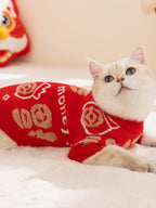 A white cat lying down, wearing a red knit sweater with gold Chinese character designs.