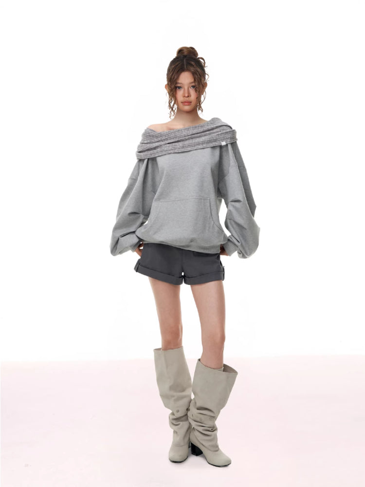 Full body shot of a model wearing a grey off-the-shoulder sweatshirt with a ribbed neckline and voluminous sleeves, grey shorts, and light-colored knee-high boots.